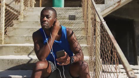 African-american-man-putting-earphones-on,-holding-smartphone,-taking-break-in-exercise-outdoors