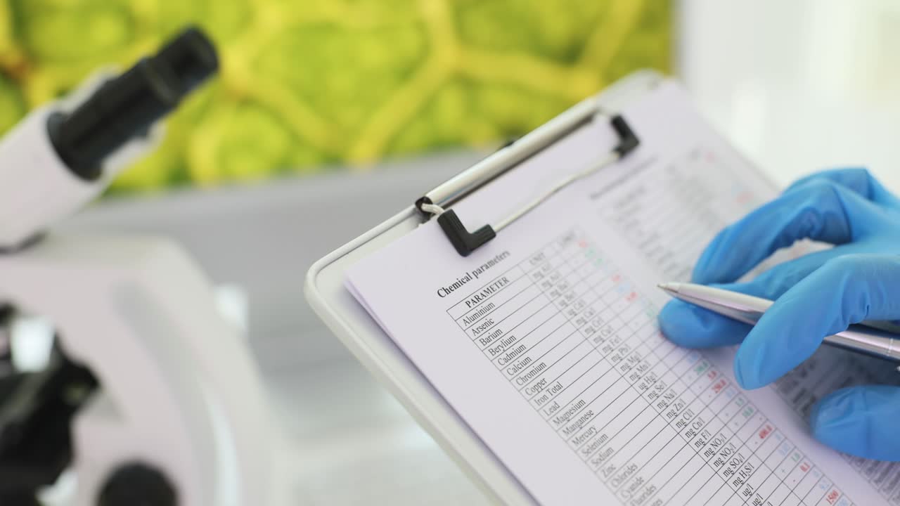 Person in lab glove analyzing chemical parameters on a clipboard next to a microscope