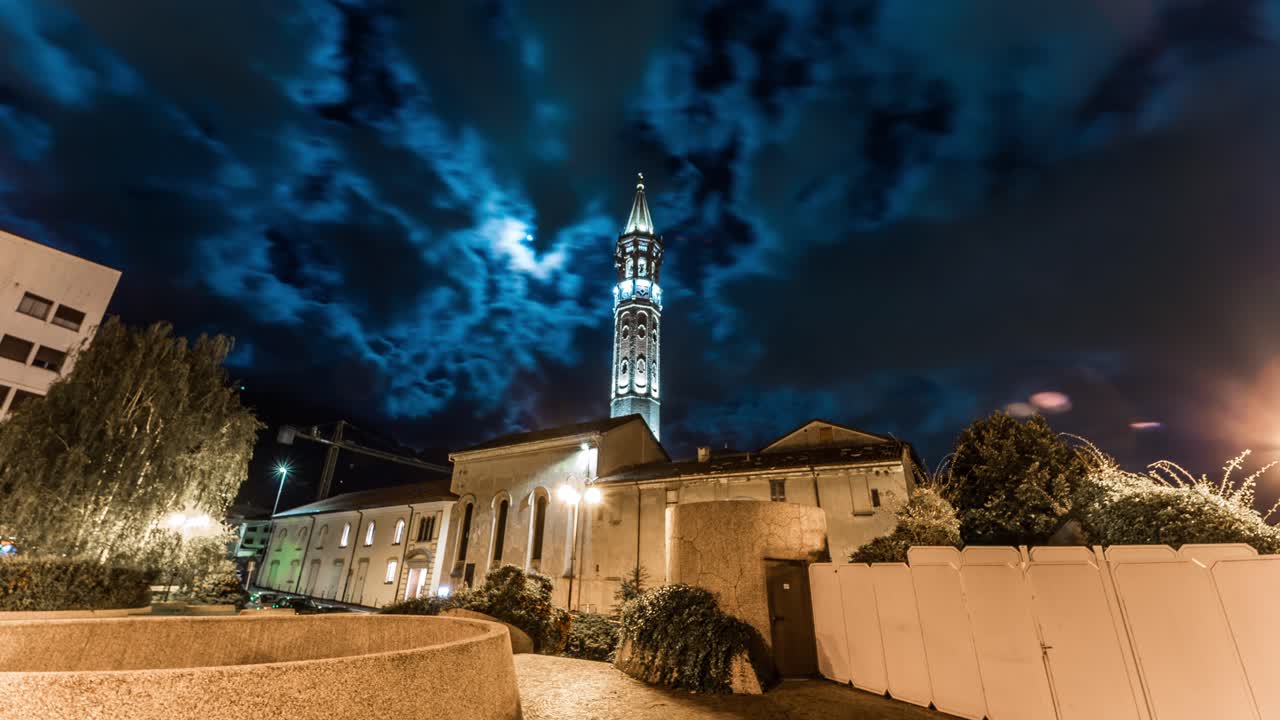 vista en timelapse de una torre de la iglesia iluminada contra un dramático cielo nocturno lleno de nubes, rodeado de varios edificios y una valla en primer plano que proporciona una atmósfera serena
