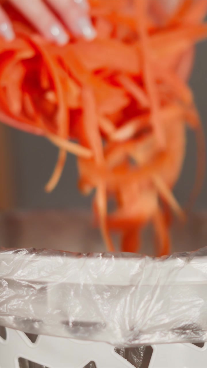 Le mani di una donna versano ripetutamente sottili bastoncini di carota arancioni in un contenitore di plastica bianco rivestito con pellicola trasparente. L'azione è costante e continua, concentrandosi sul trasferimento di scarti alimentari all'interno