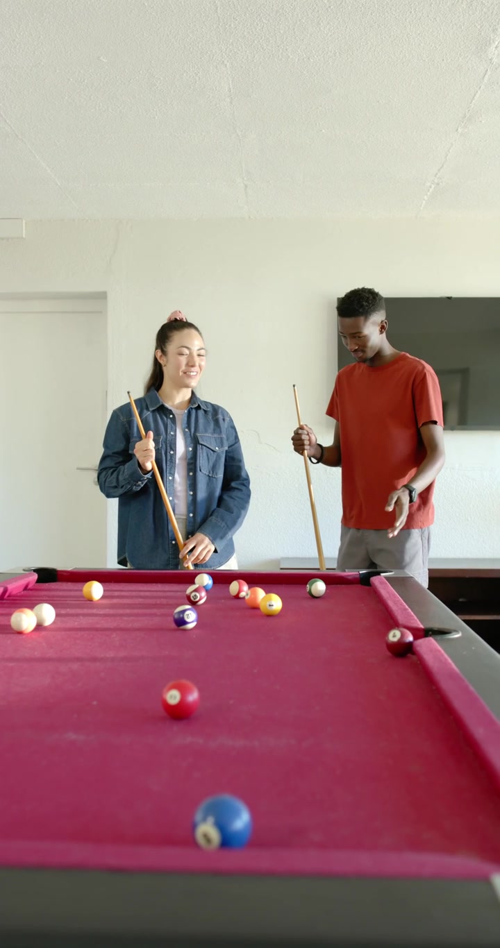 Vertical video: Couple standing behind red pool table with cues, noting balls at home, aiming shot