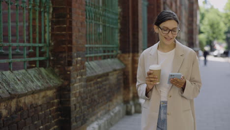 woman walking and using phone while enjoying coffee in city