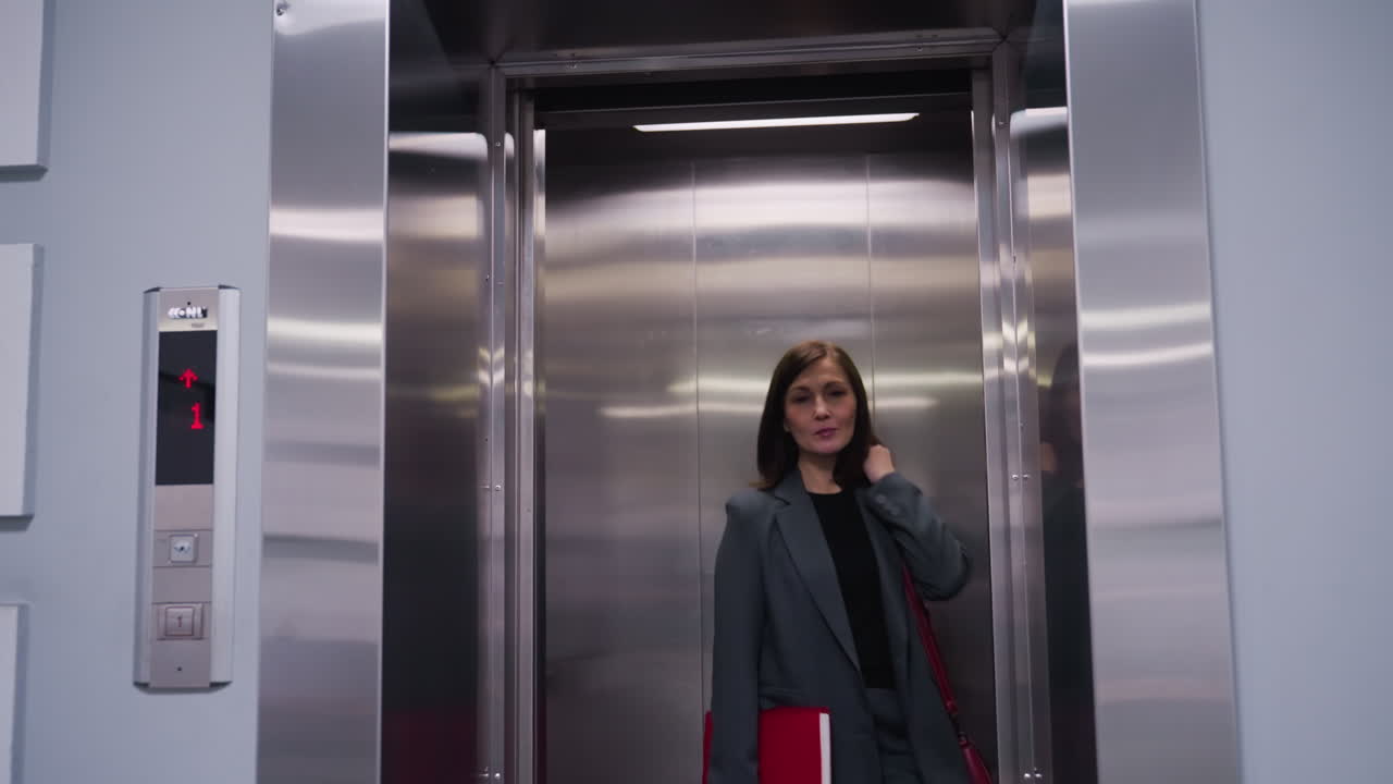 Businesswoman standing inside modern elevator with metallic walls, facing forward, ready to exit. She carries a folder and has confident posture, dressed in professional attire for corporate setting