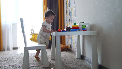 small kid moves the chairs from the table. toddler touches the toys on the desk and then lowers to the floor.