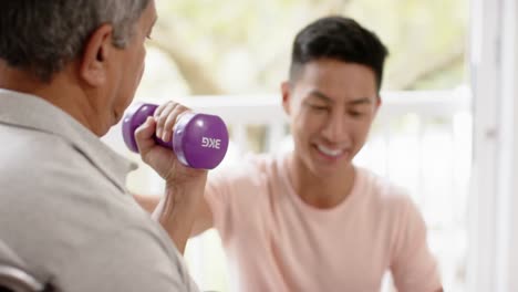 Diverse-male-physiotherapist-advising-and-senior-man-in-wheelchair-using-dumbbells,-in-slow-motion