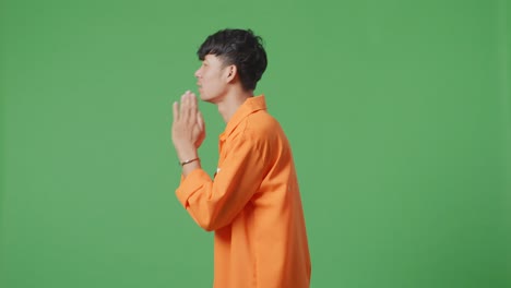 녹색 스크린 배경에 서서 무언가를 요청하는 기도 손 제스처를 하는 수갑을 입은 아시아 남성 수감자의 측면 시각