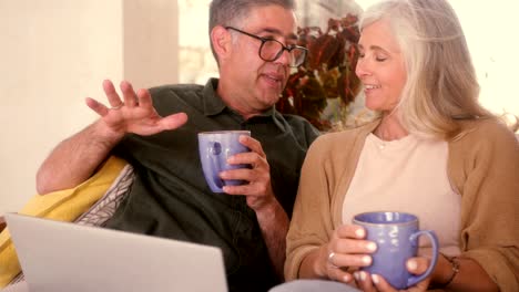 una pareja de mayores usando una computadora portátil y relajándose con café en el sofá