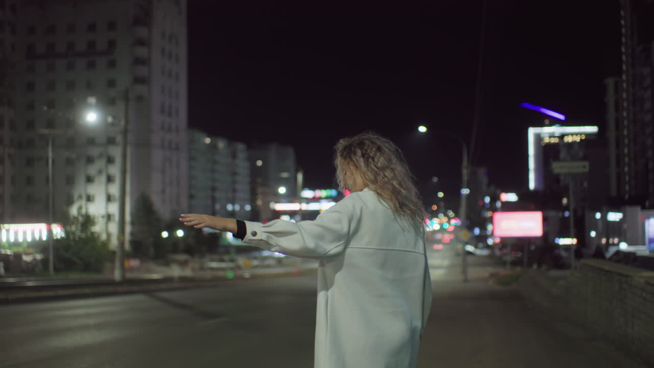Young woman in white coat stretches arm to stop cab on quiet city street at night, surrounded by colorful bokeh lights of moving cars and distant blurred view of tall residential buildings