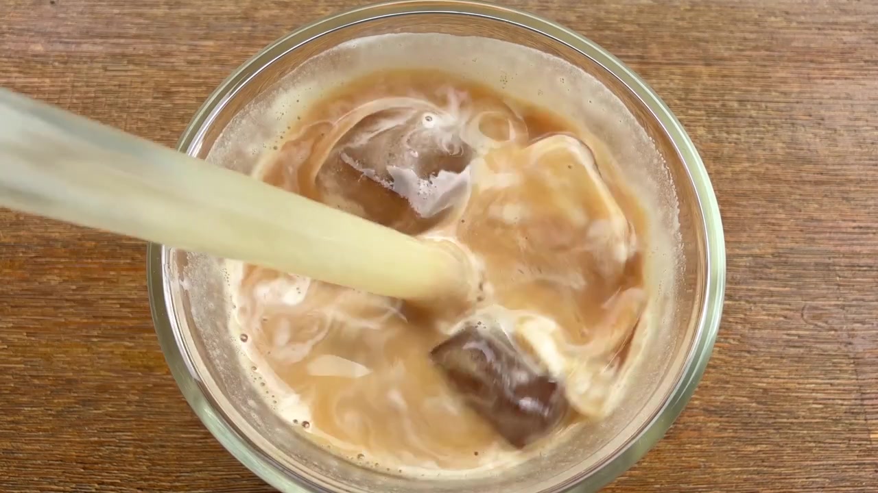 Mescolare un caffè latte ghiacciato con cannuccia di vetro e cubetti di ghiaccio, primo piano.