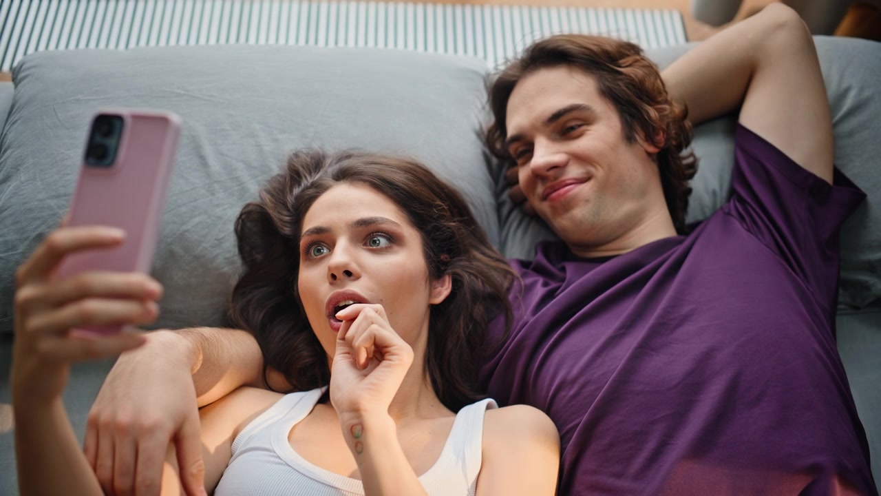 Online sweethearts relaxing mattress looking smartphone house top view closeup