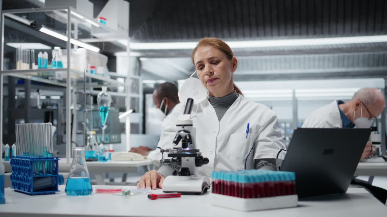 Biolog iført mundbind i medicinsk laboratorium bruger videnskabeligt mikroskop