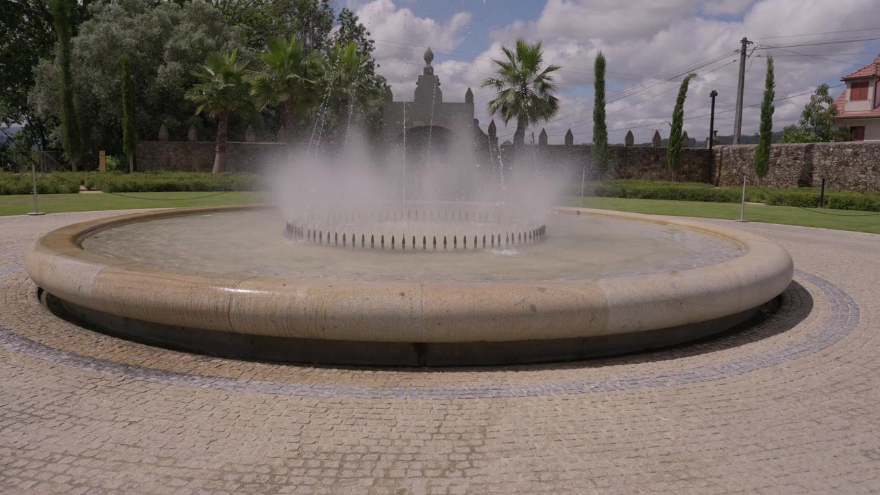 Fuente de piedra circular con chorros de agua nebulizada en una plaza adoquinada con palmeras y una capilla, Portugal.