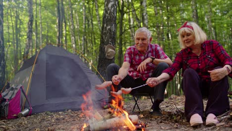 Happy-senior-man-and-woman-talking,-cooking-frying-sausages-over-campfire-in-evening-wood,-camping