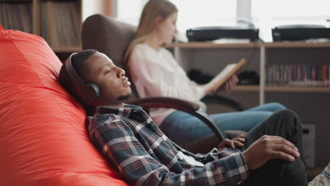 hombre escucha información de audio a través de auriculares en la biblioteca. tipo negro con auriculares se sienta en una bolsa de frijoles suave descansando en una acogedora librería. ocio nerd