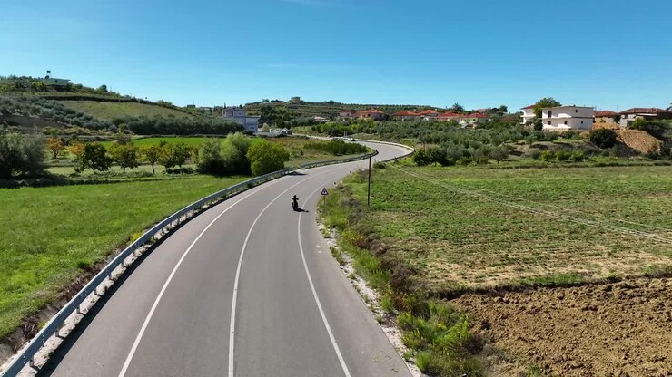 Motocicletta in corsa su strada di campagna deserta vicino a case e campi in una giornata di sole, ripresa aerea 4K 60fps