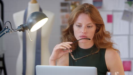 mujer cansada dueña de un negocio de moda con una copa de vino trabajando hasta tarde en la computadora portátil en el estudio