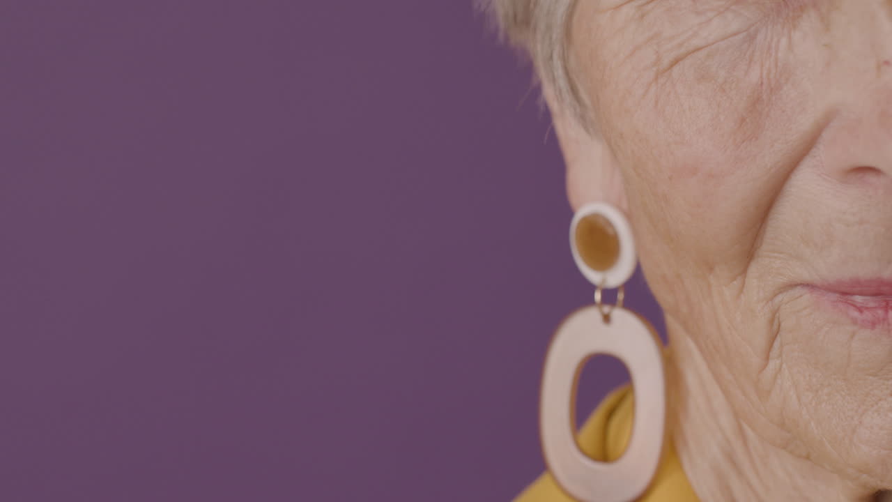 vista de cerca de la mitad de la cara de una mujer mayor con cabello corto y ojos verdes usando aretes posando con una cámara de expresión seria sobre fondo púrpura