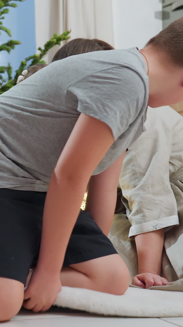 Madre e hija blancas sentadas cerca de un árbol, niña pequeña con vestido sosteniendo luces de hadas mientras el niño se arrodilla y acaricia al perro que descansa cerca, luz natural cálida, alfombra acogedora, conversación agradable, preparativos íntimos para las fiestas