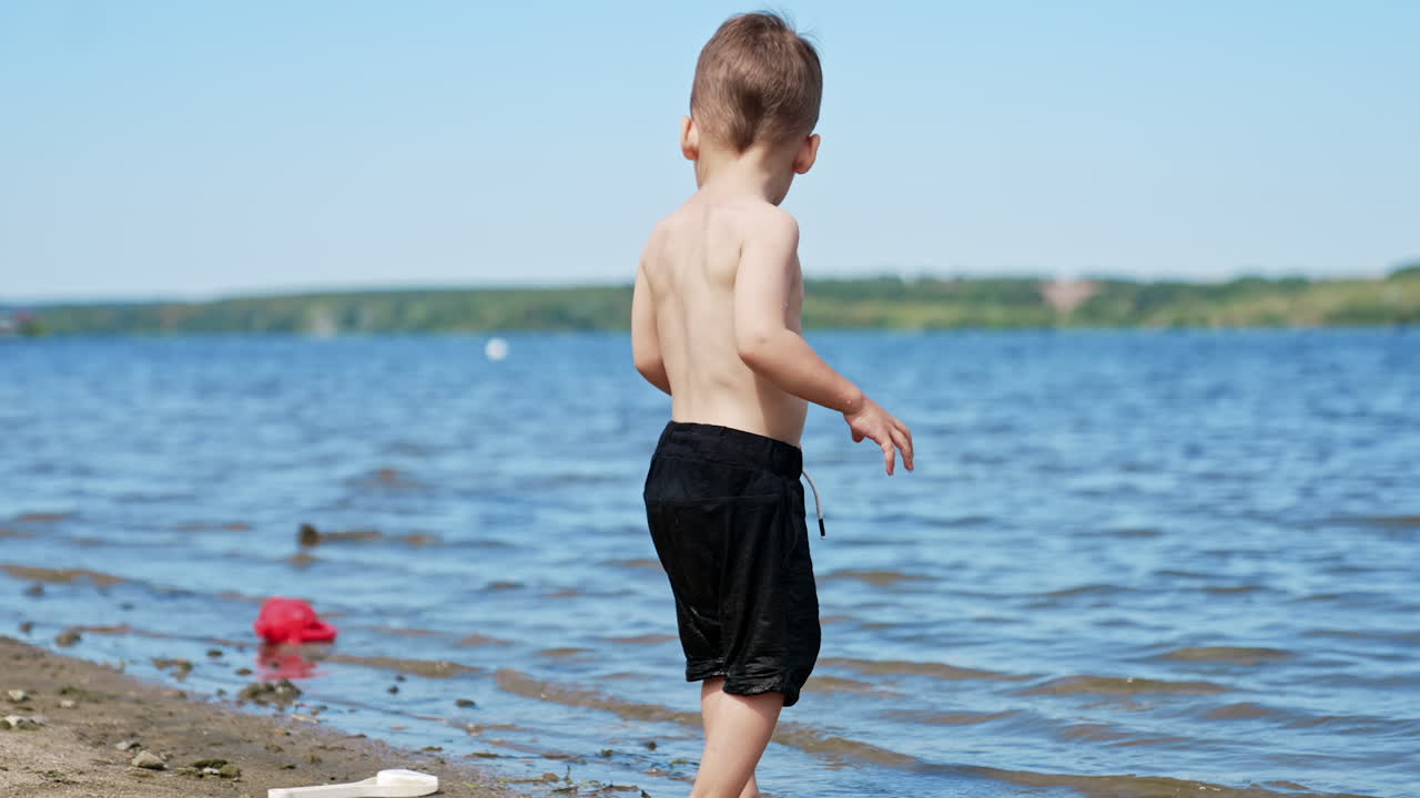Cute kid in black shorts carefully steps by the water and sand in the beach. Caucasian child having rest near the river in summer.