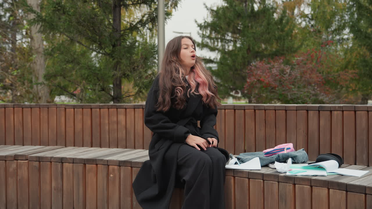 Giovane studentessa caucasica seduta su una panchina che ordina appunti di studio in un campus autunnale, cappotto lungo e zaino, cancelleria e quaderno sparsi, controlla il telefono, si sistema i capelli, momenti di contemplazione e quiete