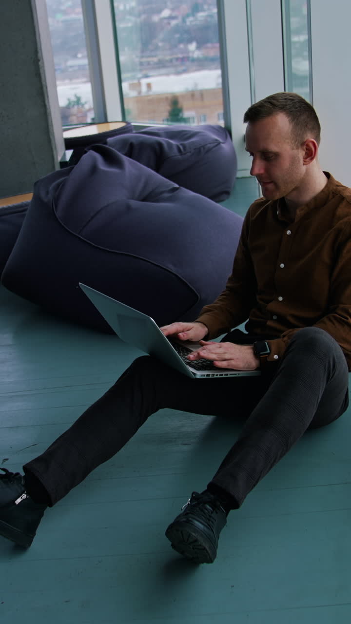 Young man with laptop sitting on floor by the window Vertical video