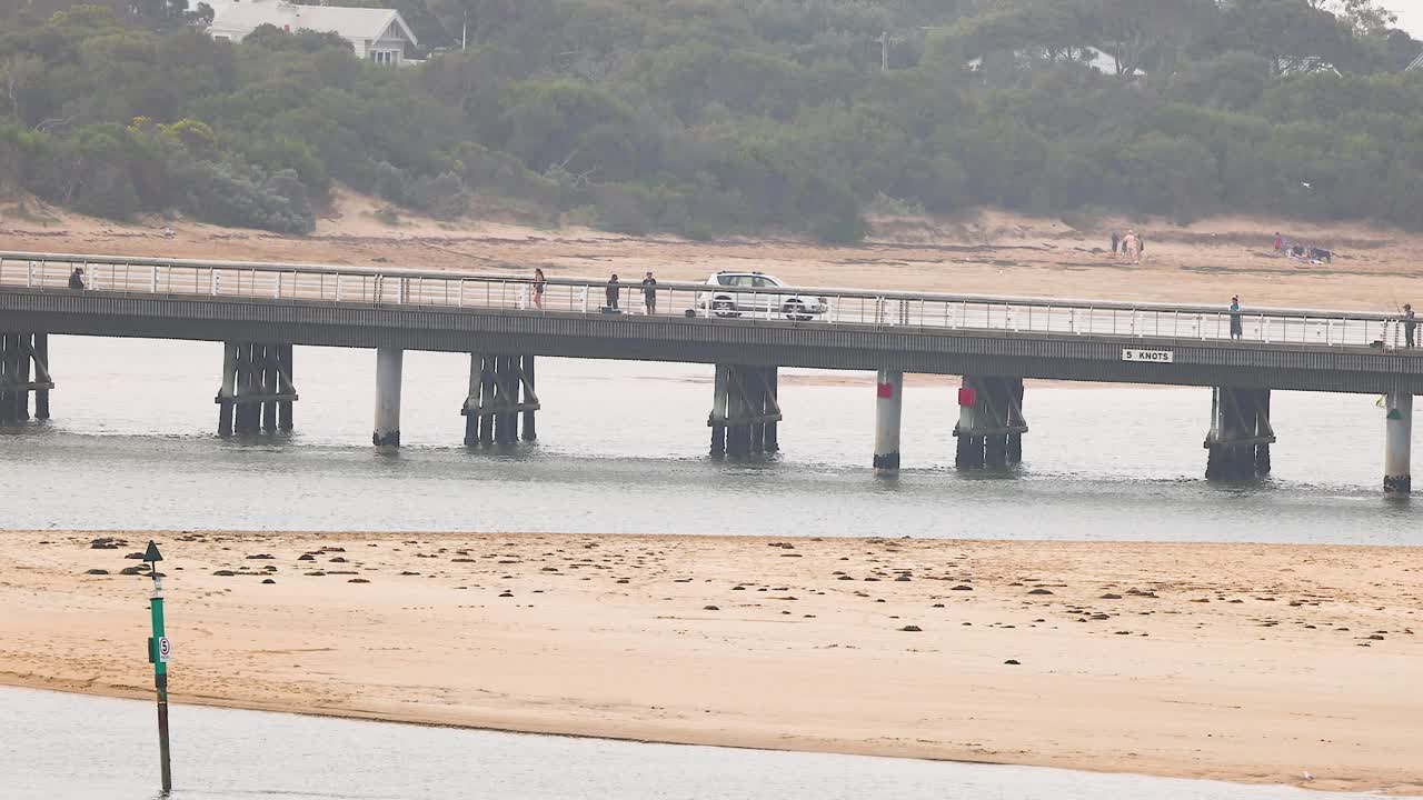 Cars and people traverse a bridge in Barwon Heads, Australia, under overcast skies. The scene captures movement and daily life