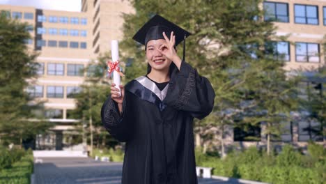 asian woman student graduates in cap and gown with diploma showing ok hand sign over eye and smiling to camera in front of a magnificent university building