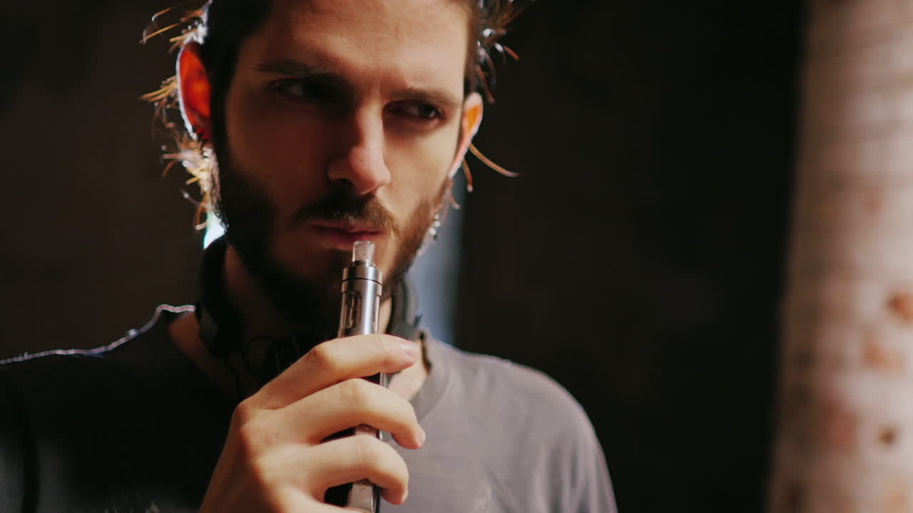 el joven hipster fuma un cigarrillo electrónico y produce vapor en un haz de luz lento