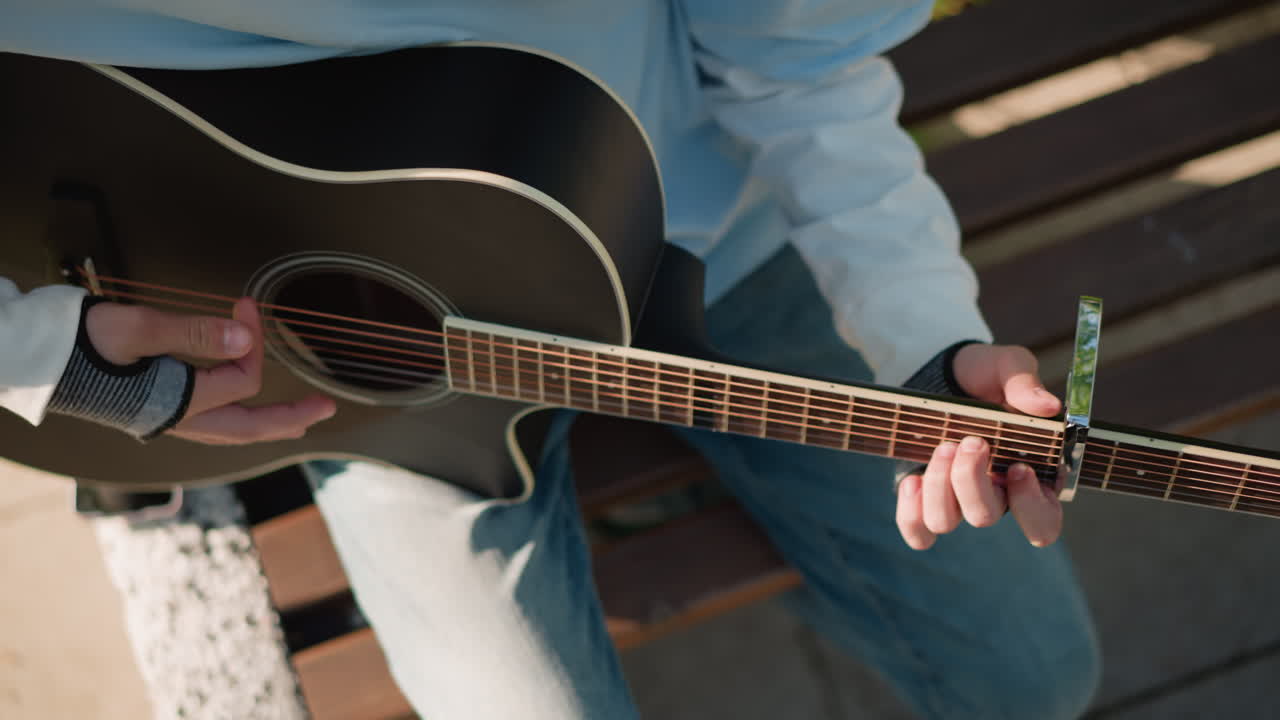 closeup hands fretting acoustic guitar capo, precision fingerstyle technique and capo placement, denim jeans on bench, sunlight highlights on wood grain, practicing chord changes with focused