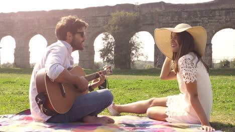 feliz joven pareja encantadora sentada acostada en una manta en la hierba frente a las ruinas del antiguo acueducto romano en el parco degli aquedotti parque romántico en roma tocar la guitarra cantar hermosa chica con sombrero bucólica cámara lenta
