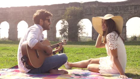 happy young lovely couple sitting lying on a blanket in the grass in front of ancient roman aqueduct ruins in parco degli acquedotti park in rome romantic play guitar sing beautiful girl with hat bucolic slow motion