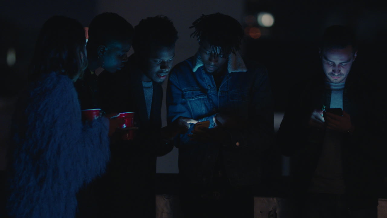 한 무리의 친구들이 밤에 지붕에서 휴식을 취하고 스마트폰 기술을 사용하여 문자 메시지를 보내고 소셜 미디어 라이프 스타일을 공유하고 온라인을 탐색하고 주말 모임을 즐기고 있습니다.