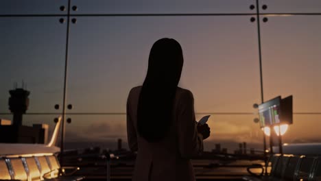 back view of asian businesswoman with rolling suitcase in boarding lounge of airline hub, waiting for flight looks at the ticket, airport terminal with airplane takes off outside the window