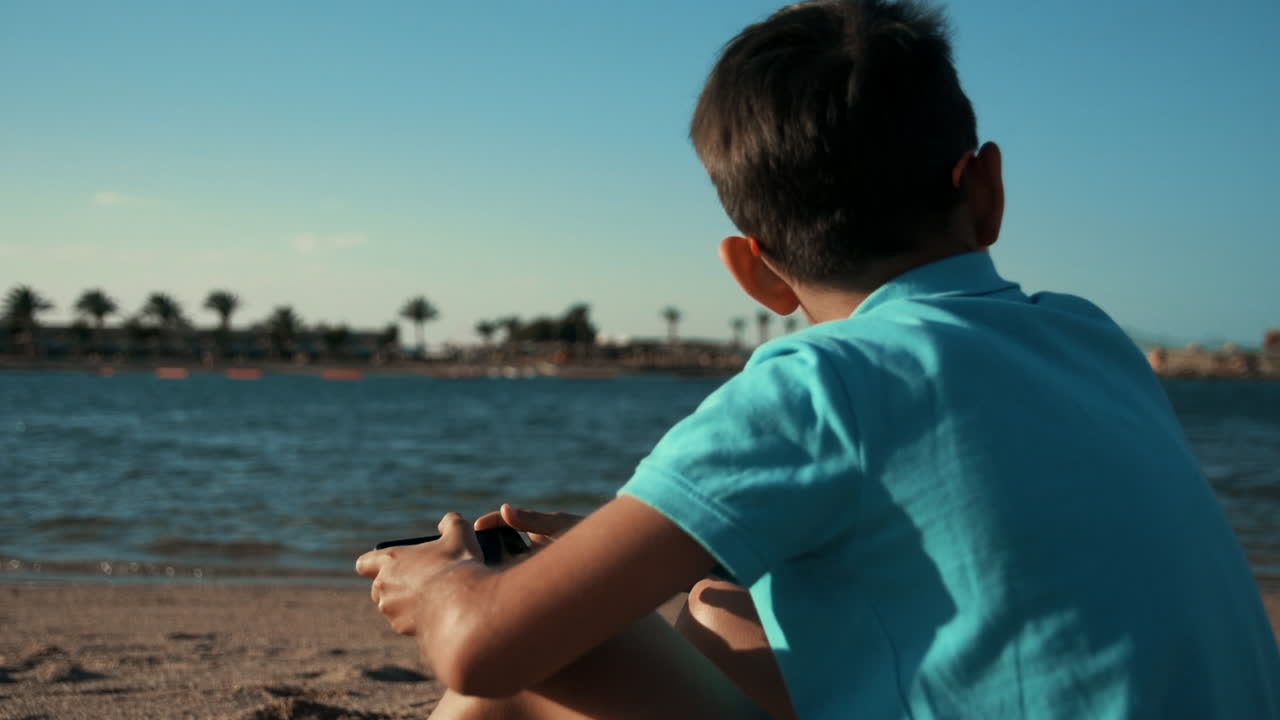 hombre joven guapo con camiseta azul sentado con teléfono móvil en la bahía del mar.
