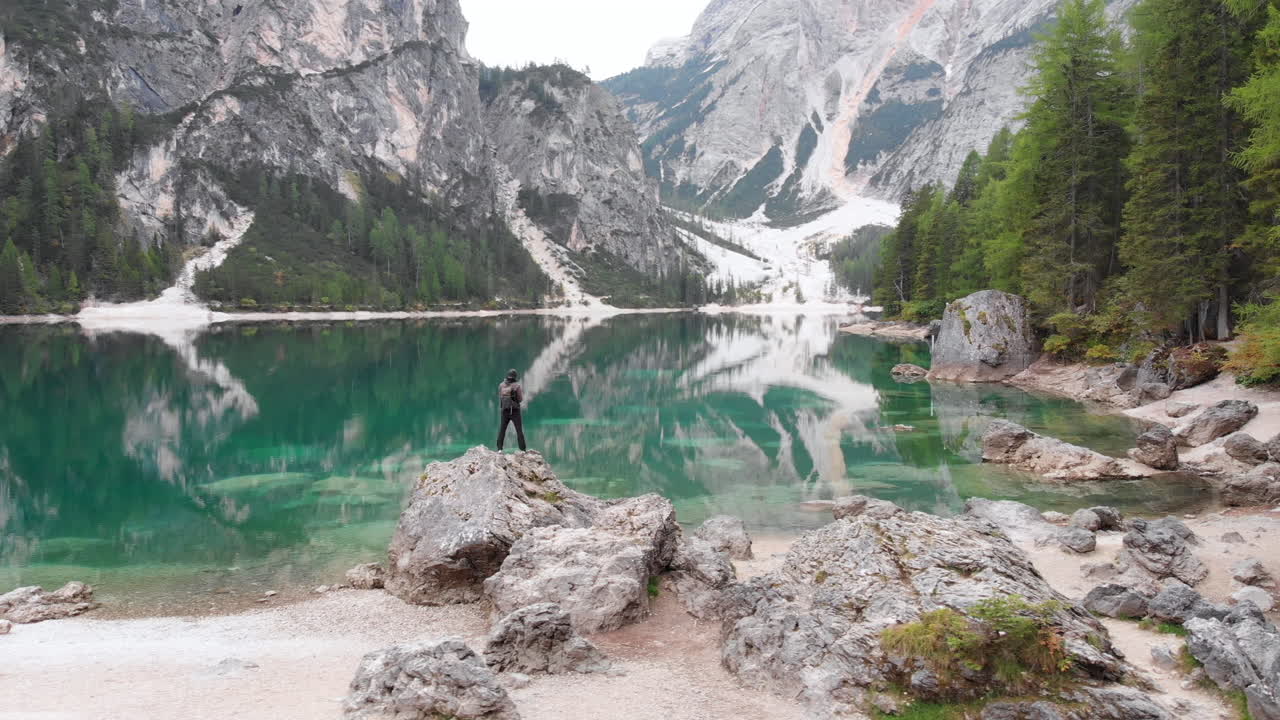 Inquadratura circolare da drone attorno a un modello uomo in piedi su una roccia che si affaccia sul lago di Braies nelle Dolomiti, Italia.