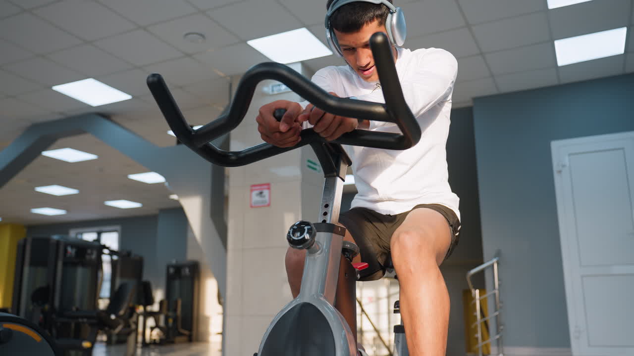 Boy in white sports top and black shorts listens to music on headphones while pedaling stationary bike in well equipped gym, with weight machines and bright lights in background