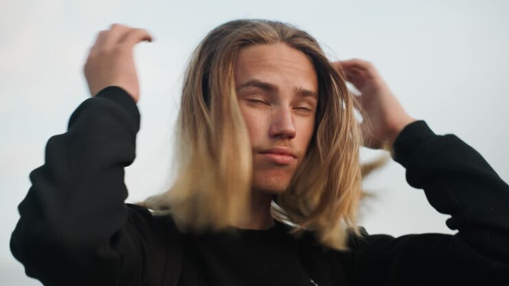 Young man adjusting hair on overpass, backpack and black sweater, soft dusk sky, contemplative gaze and gentle hand movements smoothing long hair, candid closeup portrait with natural light