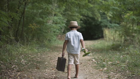 Boy-walking-in-the-forest-in-vintage-clothes-carrying-a-suitcase-and-a-toy-plane