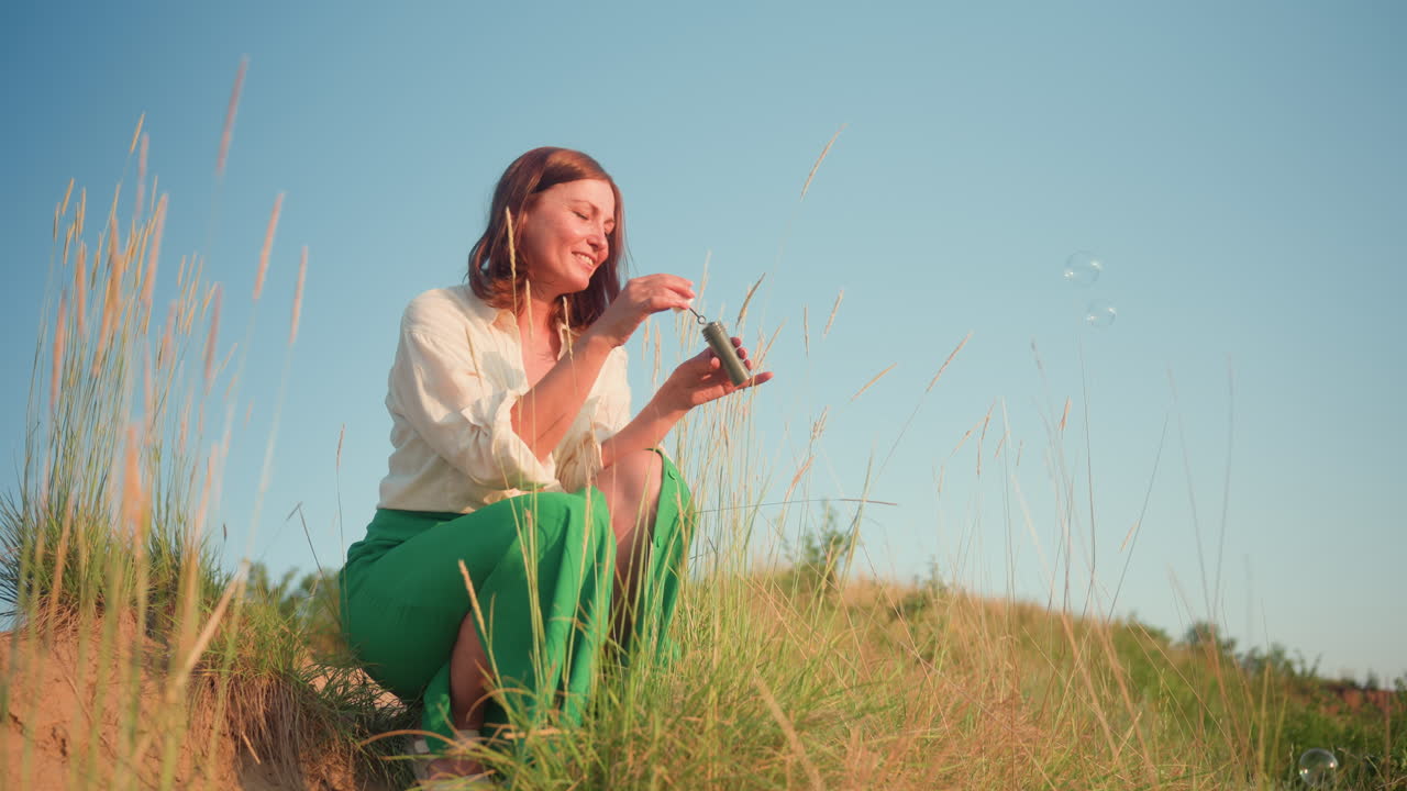 Squatting woman in green dress blowing translucent soap bubbles over grassy hillside at golden hour, watching iridescent spheres float