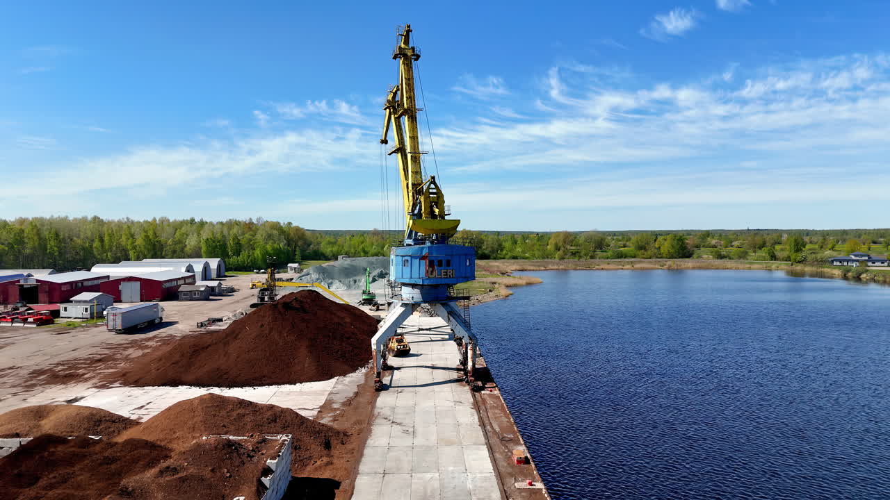 From a high drone, a bulk-handling port crane straddles a concrete quay beside calm blue water, with brown stockpiles, service buildings, and forest fringe under crisp, breezy daylight