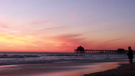 a man takes a picture of the beach during a gorgeous yellow, orange, pink and blue sunset with the huntington beach pier in the background at surf city usa california