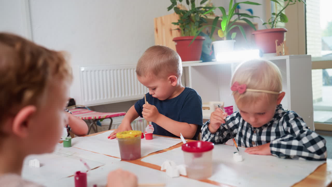 Group of children sit around table painting plaster figurines with brushes, focusing carefully on their creative task, surrounded by colorful paint containers in bright classroom environment