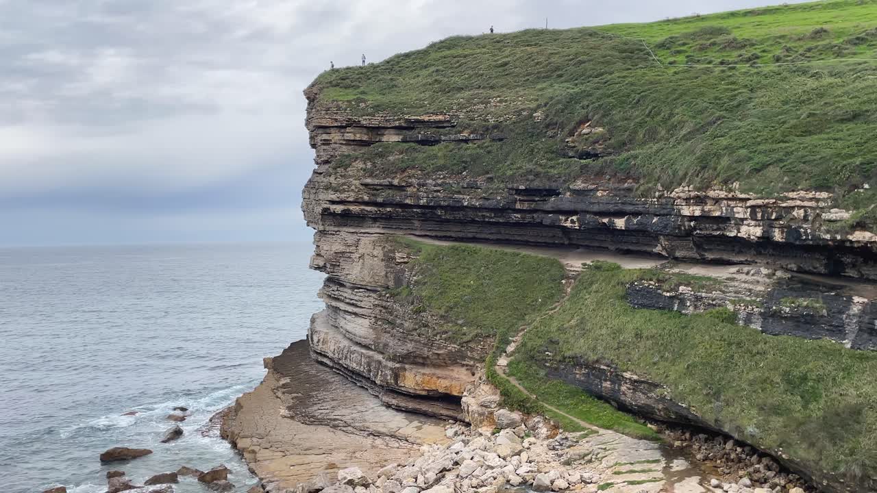 filming of the coast of the Cantabrian Sea where we see a spectacular cliff, we appreciate its stratographic layers of limestone covered by a green mantle of grass, at its top there are three people