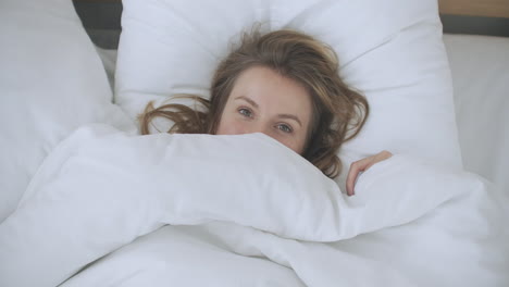 happy woman peeking out from her blanket in bed and smiling