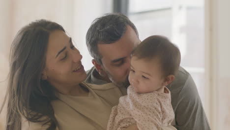 Happy-Young-Mother-Holding-Baby-Girl-In-Arms-While-Father-Coming-Up-And-Kissing-Infants-Cheek-At-Home