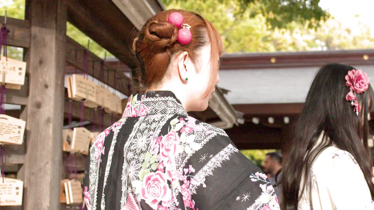 Một người phụ nữ mặc kimono họa tiết hoa đang cầu nguyện trước một bức tường gồm các thẻ cầu nguyện ema bằng gỗ.