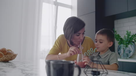 young mom and son drinking juice with candy at kitchen. happy mother spending time with son sitting on table in slow motion