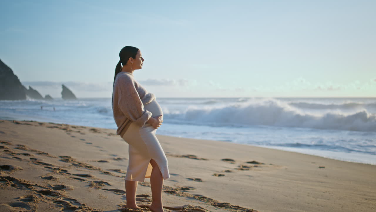 아름다운 임신한 여성이 모래 해변에서 모래 모양의 바다 파도를 상하고 있습니다.