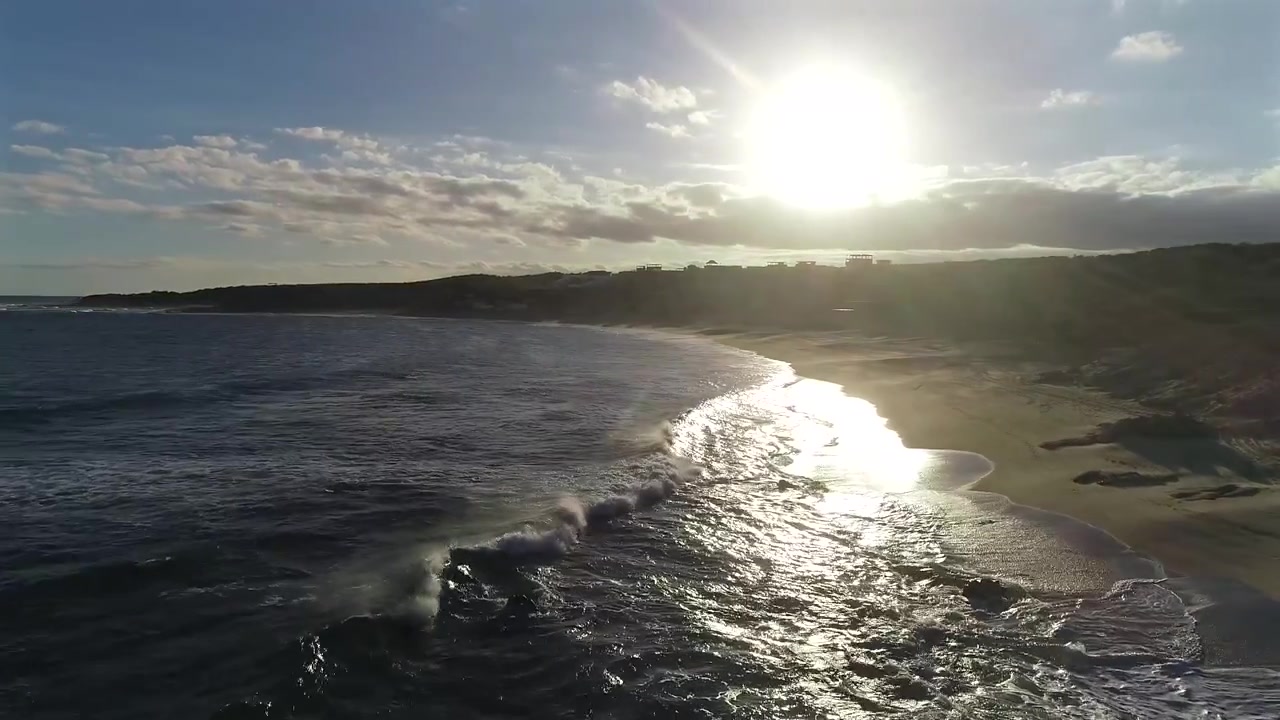 Drone flyver lavt over vandet og klipperne ved Playa El Rinconcito, drejer fra at vende mod havet til at vende mod stranden, små klitter og den sene eftermiddagssol, mens bølger ruller ind gennem hele optagelsen