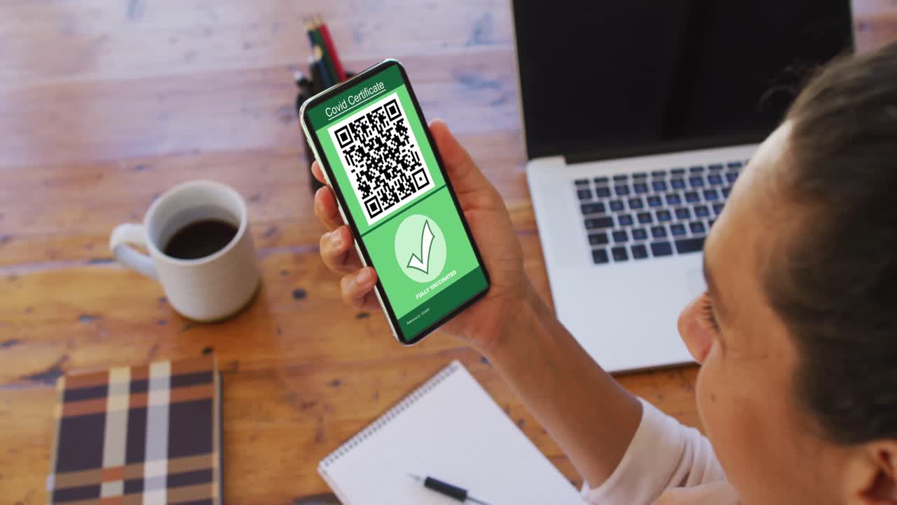 Woman at desk holding smartphone with covid vaccination certificate and qr code on screen